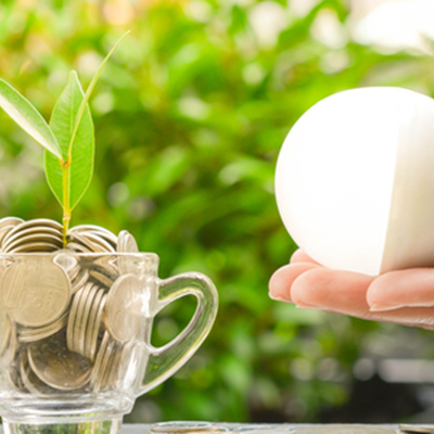 hand holding lightbuld towards cup of money with plant 